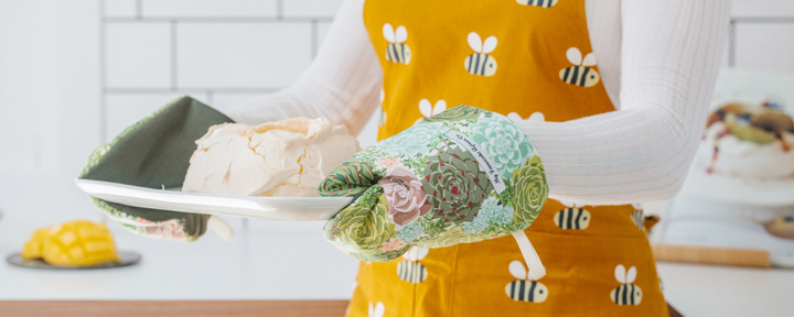 Woman holding tray of pavlova with oven mitts with succulents and and wearing a bee apron 